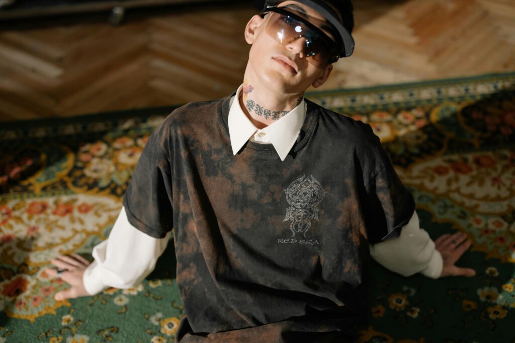 A stylish man with sunglasses and tattooed neck poses confidently indoors on a patterned rug.
